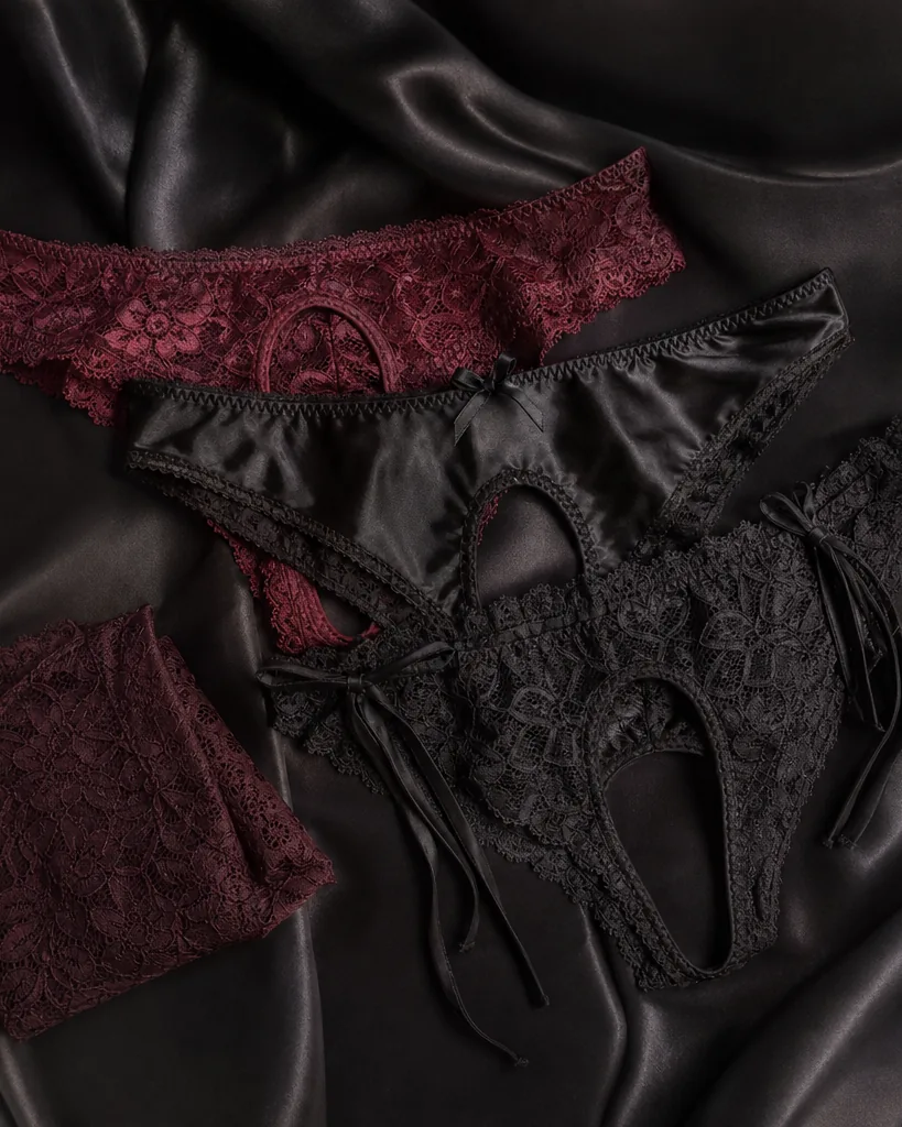 Adult woman in black lace crotchless panties and a matching bra, standing by a window in soft evening light